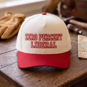May include: A white and red baseball cap with the text "ZERO PERCENT LIBERAL" embroidered in red. The cap has a red brim and a red button on top. The hat is sitting on a wooden surface.