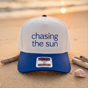 May include: A five-panel baseball cap with a cream-colored crown and a solid blue brim. The front of the crown features the text "chasing the sun" in blue lettering. A rectangular sticker with the brand name "OTTO" is centered on the brim.