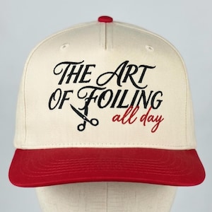May include: A baseball cap with a cream-colored crown and a red brim. The cap features the text "THE ART OF FOILING all day" in black and red script, with a scissors graphic.