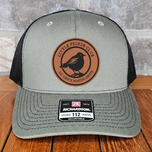 May include: A gray and black trucker hat with a brown patch featuring a bird and the text "LITTLE PECKER CLUB". The hat has a curved brim and an adjustable strap. The tag reads "RICHARDSON ORIGINAL 112 TRUCKER".