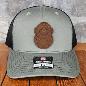 May include: A gray and black trucker hat with a brown leather patch featuring a vintage diving helmet design. The hat has a curved brim and an adjustable strap. The patch reads "RICHARDSON ORIGINAL 112 TRUCKER".