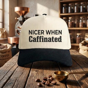 May include: A two-tone baseball cap with a cream-colored front panel and a black brim. The front panel features the black text "NICER WHEN Caffinated" printed in a bold, sans-serif font for the first line and a decorative serif font for the second.