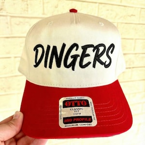 May include: A white and red baseball cap with the word "DINGERS" embroidered in black. The cap has a red brim and a small red button on top. The label reads "OTTO CLASSIC FIT OSFM MID PROFILE MAXIMUM COMFORT".