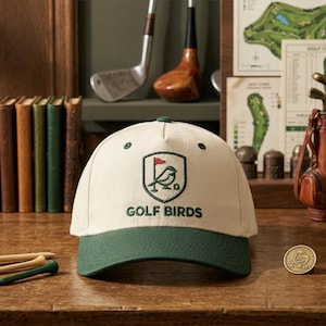 May include: A cream-colored baseball cap with a green brim and embroidered "Golf Birds" logo. The logo features a bird with a golf flag. The cap is on a wooden surface with golf tees and a golf ball marker.