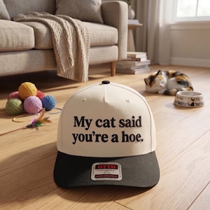 May include: A cream and black baseball cap with the text "My cat said you're a hoe." The hat is sitting on a wooden floor. A cat, yarn balls, and a couch are in the background.