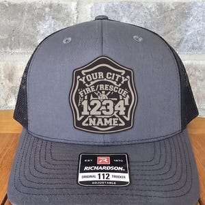 May include: A grey trucker hat with a black mesh back and a dark grey patch on the front. The patch features a fire rescue shield design with the text YOUR CITY, FIRE/RESCUE, 1234, and NAME. A black Richardson 112 Original sticker is on the brim.
