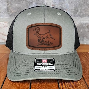 May include: A gray and black trucker hat with a brown leather patch featuring a deer leaping over a log. The hat has a curved brim and an adjustable strap. The patch has the text "RICHARDSON 112 TRUCKER" on the bottom.
