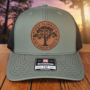 May include: A gray trucker hat with a black mesh back. The front features a brown circular patch with a tree climber design and the text "TREE CLIMBER NOT AFRAID OF HEIGHTS." The hat includes a Richardson 112 tag.