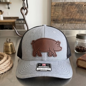 May include: A heather gray trucker hat with a black mesh back and a brown leather patch featuring a pig silhouette. The front of the brim displays a black and white sticker with the text EST. 1918 RICHARDSON 112 TRUCKER ADJUSTABLE.