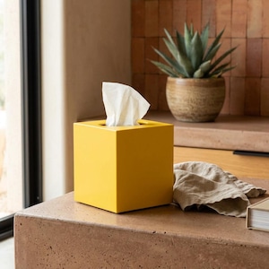 May include: A square, yellow tissue box with a white tissue sticking out of the top. The tissue box is sitting on a brown surface. A potted plant and a folded cloth are in the background.