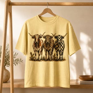 Pode incluir: Uma t-shirt de manga curta amarelo-p&aacute;lido com uma impress&atilde;o gr&aacute;fica de tr&ecirc;s bovinos longhorn lado a lado. A vaca da esquerda tem uma pelagem castanha salpicada, a do centro um padr&atilde;o de leopardo e a da direita um padr&atilde;o de xadrez preto e branco.