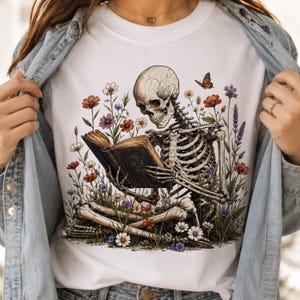 May include: A white t-shirt featuring a graphic illustration of a human skeleton sitting cross-legged while reading an open book. The skeleton is surrounded by a variety of colorful wildflowers and a single orange butterfly in a garden setting.