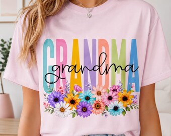 Pode incluir: Camiseta rosa claro com a palavra "GRANDMA" em letras coloridas e uma fonte cursiva. Um arranjo floral com flores roxas, amarelas e rosas adorna a parte inferior.