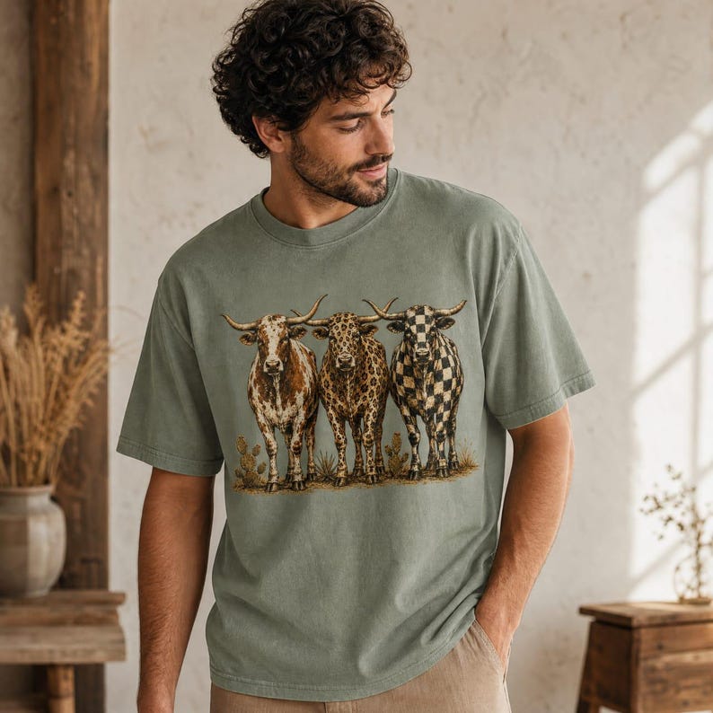 Pode incluir: Uma t-shirt de manga curta verde s&aacute;lvia com uma impress&atilde;o gr&aacute;fica de tr&ecirc;s bovinos longhorn num cen&aacute;rio des&eacute;rtico. Os bovinos t&ecirc;m padr&otilde;es distintos: um &eacute; castanho e branco sarapintado, um tem padr&atilde;o de leopardo e um &eacute; axadrezado a preto e branco.