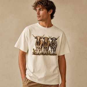 Pode incluir: Uma t-shirt de manga curta cor de creme com uma impress&atilde;o gr&aacute;fica de tr&ecirc;s bovinos longhorn de p&eacute; juntos. Os bovinos t&ecirc;m padr&otilde;es distintos, incluindo manchas, estampado de leopardo e um design de xadrez preto e branco com pequenos cactos.