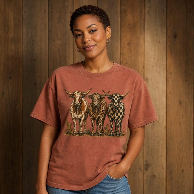 Pode incluir: Uma t-shirt de manga curta cor de terracota com uma impress&atilde;o gr&aacute;fica de tr&ecirc;s bovinos longhorn lado a lado. Os bovinos t&ecirc;m padr&otilde;es distintos: um &eacute; salpicado, um tem padr&atilde;o de leopardo e um tem um padr&atilde;o de xadrez preto e branco.