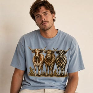 Pode incluir: Uma t-shirt de manga curta azul-clara com uma estampa gr&aacute;fica de tr&ecirc;s bovinos longhorn num cen&aacute;rio des&eacute;rtico. Os bovinos t&ecirc;m padr&otilde;es distintos: um &eacute; castanho e branco, um tem padr&atilde;o de leopardo e um tem um padr&atilde;o de xadrez preto e branco.