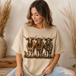 Pode incluir: Uma t-shirt bege de manga curta com uma impress&atilde;o gr&aacute;fica de tr&ecirc;s bovinos longhorn de p&eacute; lado a lado. Os bovinos t&ecirc;m padr&otilde;es distintos, incluindo manchas castanhas, estampado de leopardo e um padr&atilde;o de xadrez preto e branco.