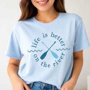 Könnte beinhalten: Hellblaues T-Shirt mit dem Schriftzug "life is better on the river" in einem kreisförmigen Design. Das Design umfasst gekreuzte Ruder und Wellengrafiken in Blautönen. Ein lässiges, bequemes T-Shirt.