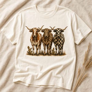 Pode incluir: Uma t-shirt de cor creme com uma impress&atilde;o gr&aacute;fica de tr&ecirc;s bovinos longhorn num cen&aacute;rio des&eacute;rtico. A primeira vaca tem uma pelagem castanha salpicada, a segunda um padr&atilde;o de leopardo e a terceira um design de xadrez preto e branco.