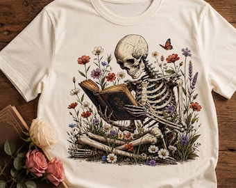 Pode incluir: Uma t-shirt de cor creme com uma impressão gráfica de um esqueleto humano sentado de pernas cruzadas enquanto lê um livro aberto. O esqueleto está rodeado por uma variedade de flores silvestres coloridas e uma borboleta laranja acima.