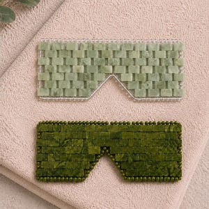 Op de afbeelding: Twee rechthoekige oogmaskers opgebouwd uit kleine, gepolijste steentjes. Het bovenste masker heeft licht saliegroene steentjes, terwijl het onderste masker bestaat uit donker olijfgroene steentjes. Beide maskers hebben een neusuitsparing.