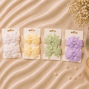 Könnte beinhalten: Vier Sets mit je zwei Stoffschleifen auf weißen, rechteckigen Präsentationskarten. Die Sets sind in den Farben Weiß, Hellgelb, Salbeigrün und Lavendel gehalten. Jede Schleife hat ein klassisches Design mit zwei Schlaufen und zwei Enden.