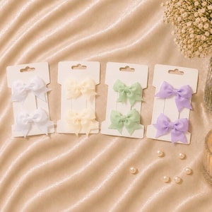 May include: Four white rectangular cards each holding two small fabric bows. The bows are arranged in pairs on each card, featuring soft pastel colors including white, cream, light green, and lavender. Each card has a cutout at the top for hanging.