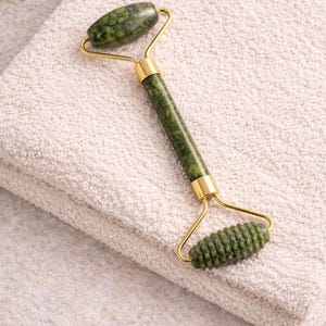 Könnte beinhalten: Eine grüne Gesichtsrolle aus Jade mit goldfarbenen Metallakzenten liegt auf einem gefalteten weißen Handtuch. Die Rolle hat zwei Jadessteine, einen ovalen und einen zylindrischen, die durch einen goldfarbenen Rahmen und Griff verbunden sind. Der Jade hat ein gesprenkeltes Muster.