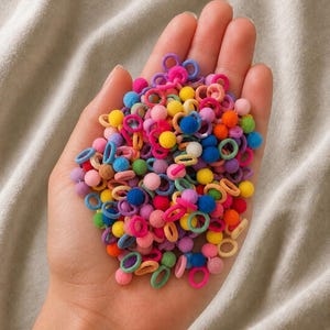 Könnte beinhalten: Eine Handvoll bunter Haargummis und kleiner Pompons in verschiedenen Rosa-, Blau-, Gelb-, Grün- und Orangetönen. Die Haaraccessoires werden in einer offenen Hand vor einem neutralen Hintergrund gehalten.
