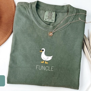 May include: A moss green Comfort Colors t-shirt with a white duck wearing sunglasses and the word "FUNCLE" printed below. A gold necklace and a brown hat are next to the shirt. The shirt is folded on a white surface.