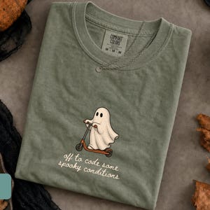 May include: A moss green t-shirt featuring a graphic of a white ghost riding a red scooter. Below the graphic, the text reads, "off to code some spooky conditions" in a white script font. The shirt has a Comfort Colors brand label at the neck.