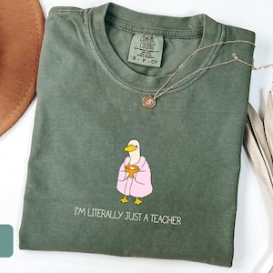 May include: Moss green t-shirt with a duck graphic wearing a pink robe and holding a golden chalice. The text "I'M LITERALLY JUST A TEACHER" is printed below the graphic. A gold necklace is draped across the shirt.
