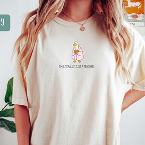 May include: Ivory-colored t-shirt featuring a cartoon duck wearing a pink robe and holding a golden chalice. The text "I'M LITERALLY JUST A TEACHER" is printed below the duck. The shirt has a relaxed fit.