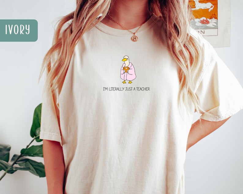May include: Ivory-colored t-shirt featuring a cartoon duck wearing a pink robe and holding a golden chalice. The text "I'M LITERALLY JUST A TEACHER" is printed below the duck. The shirt has a relaxed fit.