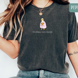 May include: Dark gray t-shirt with the text "I'M LITERALLY JUST A TEACHER". The shirt features a cartoon duck wearing a pink robe and holding a book. The shirt is a Pepper color.