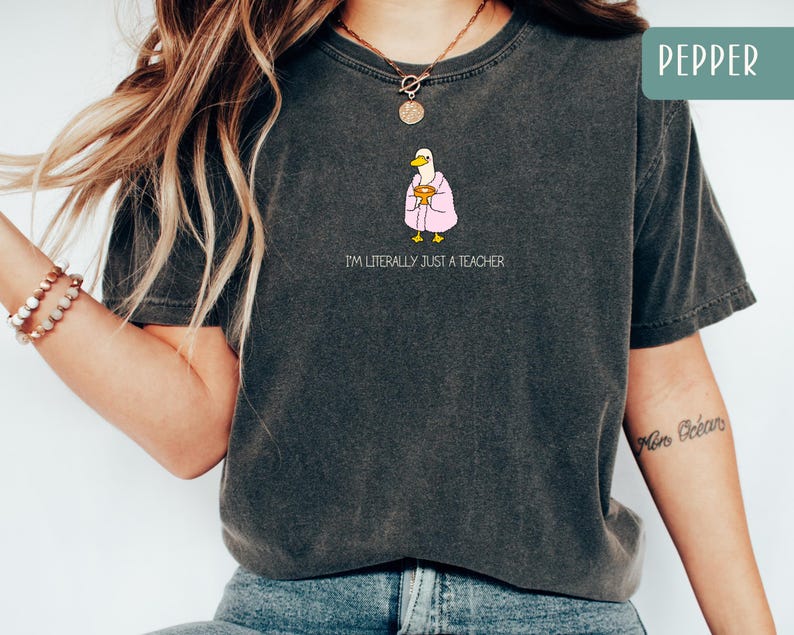 May include: Dark gray t-shirt with the text "I'M LITERALLY JUST A TEACHER". The shirt features a cartoon duck wearing a pink robe and holding a book. The shirt is a Pepper color.