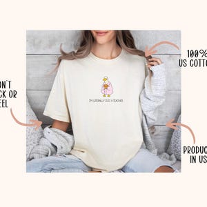 May include: Cream-colored t-shirt with a cartoon duck wearing a pink robe and holding a mug. The text "I'M LITERALLY JUST A TEACHER" is below the duck. The shirt is made of 100% US cotton.