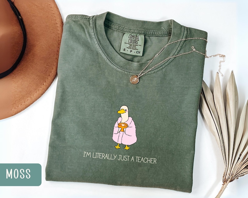 May include: Moss green t-shirt with a duck graphic wearing a pink robe and holding a golden chalice. The text "I'M LITERALLY JUST A TEACHER" is printed below the graphic. A gold necklace is draped across the shirt.