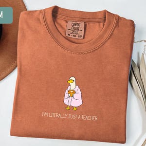 May include: A terracotta-colored t-shirt with a crew neck and short sleeves. The shirt features a cartoon duck wearing a pink robe and holding a mug. The text "I'M LITERALLY JUST A TEACHER" is printed below the duck.
