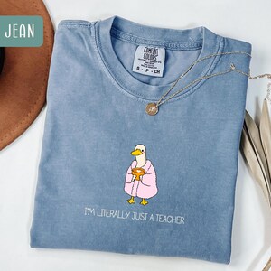 May include: Blue jean Comfort Colors t-shirt with a cartoon duck wearing a pink robe and holding a golden chalice. The text "I'M LITERALLY JUST A TEACHER" is printed below the duck. A gold necklace with a pendant is on the shirt.