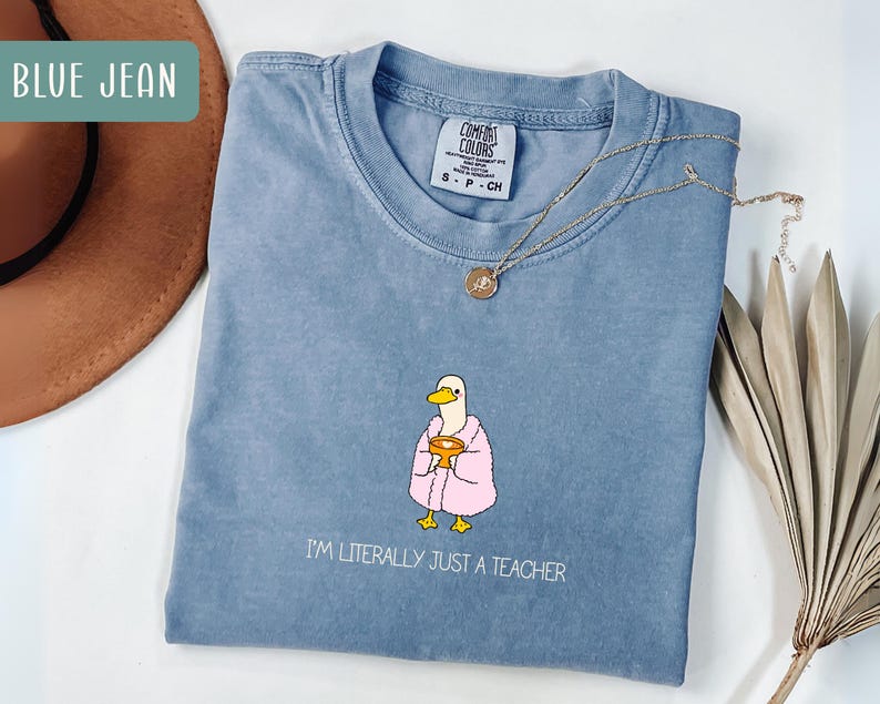 May include: Blue jean Comfort Colors t-shirt with a cartoon duck wearing a pink robe and holding a golden chalice. The text "I'M LITERALLY JUST A TEACHER" is printed below the duck. A gold necklace with a pendant is on the shirt.