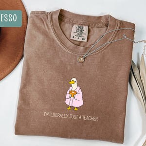 May include: Espresso-colored t-shirt with a graphic of a duck wearing a pink robe and holding a golden chalice. The text "I'M LITERALLY JUST A TEACHER" is printed below the graphic. A silver necklace is draped across the shirt.