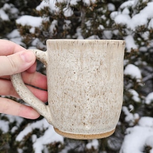 May include: A speckled beige ceramic mug with a rounded handle. The mug has a textured surface and a brown base. The background features snow-covered evergreen trees.