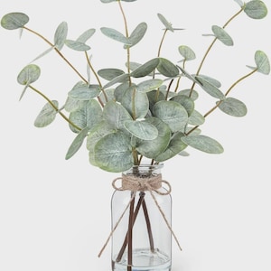 May include: A clear glass bottle vase containing a bunch of artificial eucalyptus stems with round, sage green leaves. The neck of the bottle is wrapped with a piece of natural brown twine tied into a simple bow, securing the stems inside.