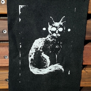 Peut inclure: Un écusson rectangulaire en tissu noir présentant un motif sérigraphié blanc représentant un chat assis. Le chat est illustré de manière stylisée et contrastée avec des détails de fourrure texturés et deux petits points blancs de chaque côté.