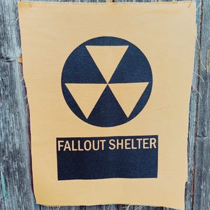 Può includere: Un cartello in tessuto beige con un simbolo di radiazione nero all'interno di un cerchio e la scritta "FALLOUT SHELTER" in un rettangolo nero. Lo sfondo è in legno grigio invecchiato.