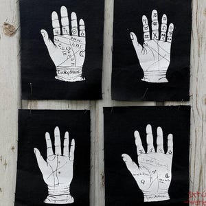 Könnte beinhalten: Vier rechteckige schwarze Stoffaufnäher mit weißen Siebdruck-Illustrationen menschlicher Handflächen. Jede Handfläche zeigt deutliche chiromantische Markierungen, einschließlich Linien, astrologischer Symbole und französischem Text.