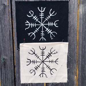 Könnte beinhalten: Zwei quadratische Stoffaufnäher mit dem Vegvisir-Symbol. Der obere Aufnäher ist schwarz mit einem weißen Druckmotiv, während der untere Aufnäher cremefarben mit einem schwarzen Druckmotiv ist. Beide Aufnäher haben rohe Kanten und sind festgesteckt.