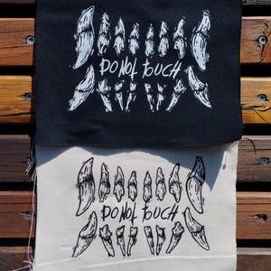 Peut inclure: Deux écussons en tissu rectangulaires, l'un noir avec de l'encre blanche et l'autre blanc cassé avec de l'encre noire. Chaque écusson présente une illustration graphique d'une rangée de dents pointues avec le texte manuscrit DO NOT TOUCH au centre.
